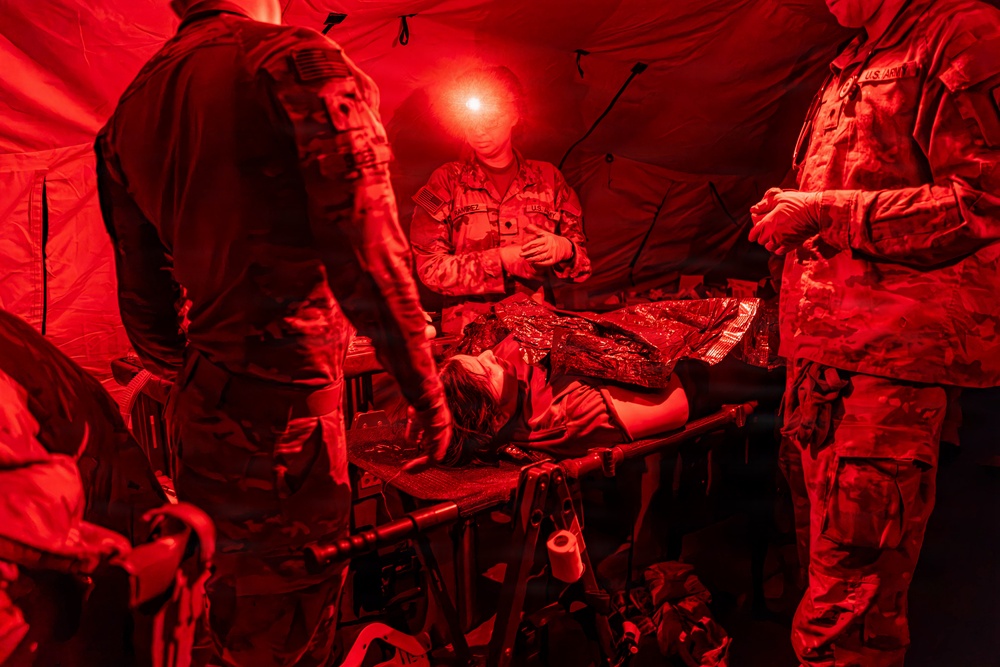 U.S. Army personnel are conducting a field medical treatment exercise inside a tactical tent illuminated by red light. A simulated casualty lies on a field stretcher/litter covered with a thermal emergency blanket while multiple soldiers attend to them. The red lighting is consistent with night operations protocols to preserve night vision.