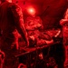 U.S. Army personnel are conducting a field medical treatment exercise inside a tactical tent illuminated by red light. A simulated casualty lies on a field stretcher/litter covered with a thermal emergency blanket while multiple soldiers attend to them. The red lighting is consistent with night operations protocols to preserve night vision.