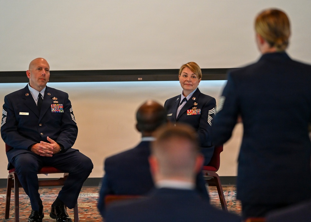 Two senior U.S. Air Force enlisted members in dress blue uniforms are seated at the front of what appears to be a formal military ceremony or briefing. They are facing an audience and a standing individual whose back is to the camera, suggesting a recognition or promotion event. Multiple uniformed personnel are visible in the foreground as attendees.