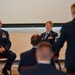 Two senior U.S. Air Force enlisted members in dress blue uniforms are seated at the front of what appears to be a formal military ceremony or briefing. They are facing an audience and a standing individual whose back is to the camera, suggesting a recognition or promotion event. Multiple uniformed personnel are visible in the foreground as attendees.