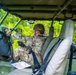 A soldier in military camouflage uniform and combat helmet is seated in the driver's seat of a military tactical vehicle, interacting with a tablet device mounted near the steering wheel. A paper map is visible on the seat or lap area, suggesting navigation or mission planning activities. The vehicle appears to be stationary in a wooded or forested environment.