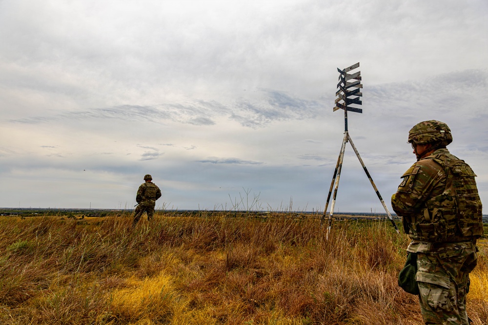 Two soldiers in camouflage uniforms and tactical gear are positioned in an open grassy field under an overcast sky. A tripod-mounted structure with a cross-shaped or multi-panel target/marker device is visible in the foreground-right. The scene appears to be a military training exercise or observation post in a flat, rural landscape.