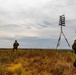 Two soldiers in camouflage uniforms and tactical gear are positioned in an open grassy field under an overcast sky. A tripod-mounted structure with a cross-shaped or multi-panel target/marker device is visible in the foreground-right. The scene appears to be a military training exercise or observation post in a flat, rural landscape.