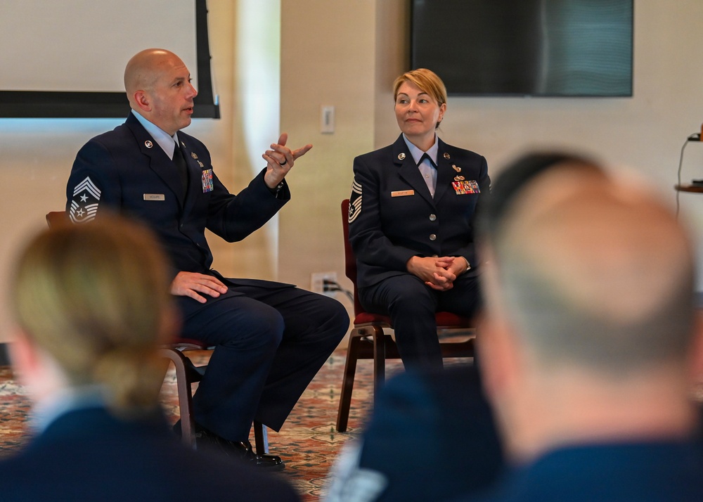 Two U.S. Air Force personnel in dress blue uniforms are seated facing an audience in what appears to be a briefing or panel discussion setting. The male on the left, wearing senior enlisted rank insignia (Chief Master Sergeant chevrons), is actively speaking and gesturing with his hand. The female on the right, wearing Master Sergeant insignia, is listening attentively with hands clasped.