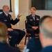 Two U.S. Air Force personnel in dress blue uniforms are seated facing an audience in what appears to be a briefing or panel discussion setting. The male on the left, wearing senior enlisted rank insignia (Chief Master Sergeant chevrons), is actively speaking and gesturing with his hand. The female on the right, wearing Master Sergeant insignia, is listening attentively with hands clasped.