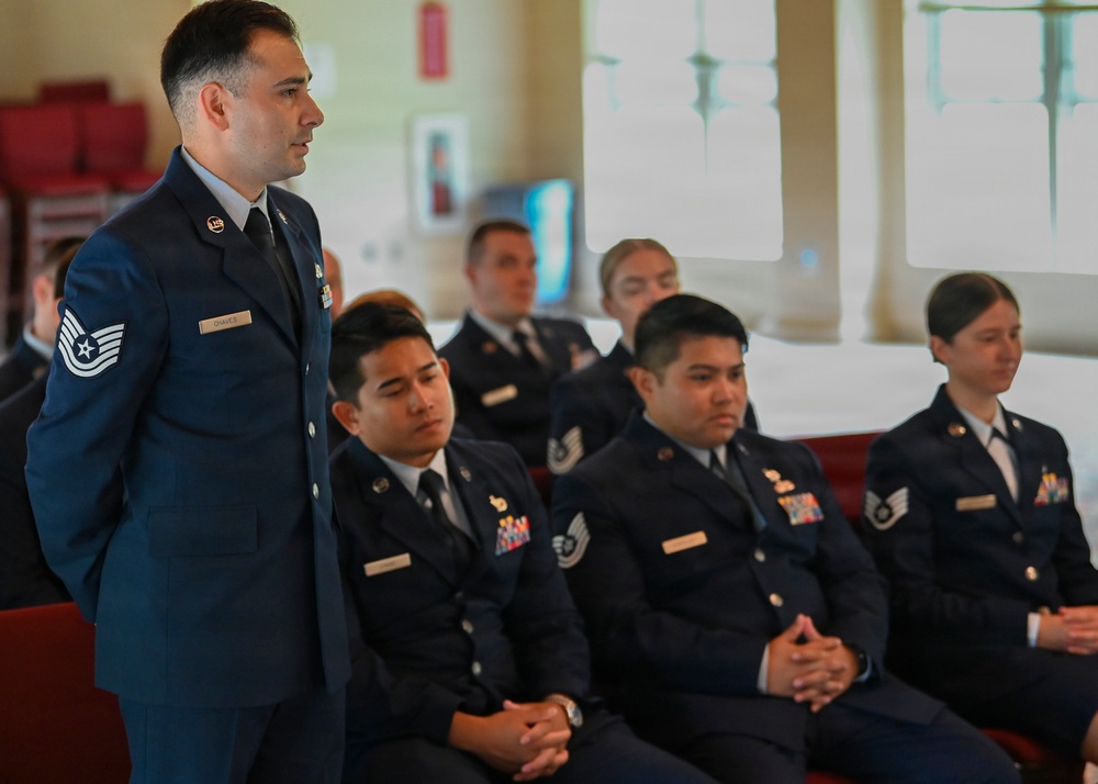A group of United States Air Force personnel in dress blue uniforms are gathered in what appears to be an indoor ceremony or formal meeting. One staff sergeant is standing while others remain seated in red chairs. The setting appears to be a chapel or formal hall based on the architectural features visible in the background.