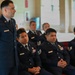 A group of United States Air Force personnel in dress blue uniforms are gathered in what appears to be an indoor ceremony or formal meeting. One staff sergeant is standing while others remain seated in red chairs. The setting appears to be a chapel or formal hall based on the architectural features visible in the background.