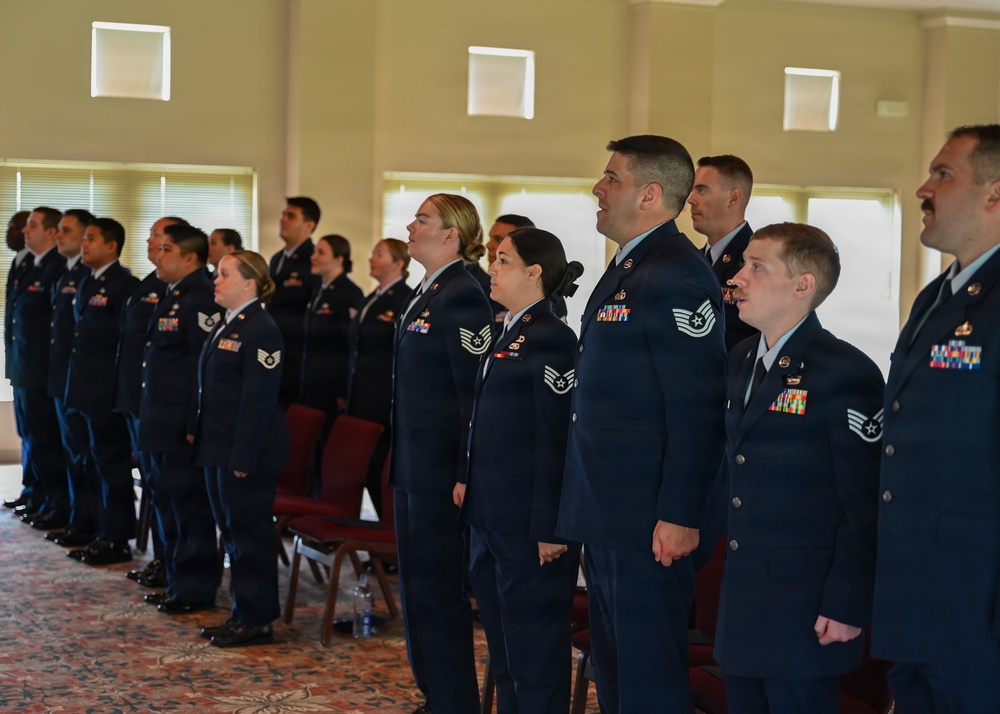 A group of United States Air Force personnel in dress blue uniforms are standing at attention in formation inside what appears to be a ceremonial hall or event venue. The service members are arranged in rows and appear to be participating in a formal military ceremony, possibly reciting an oath or singing. Both male and female airmen of various ranks are visible, identifiable by chevron insignia and service ribbons on their uniforms.