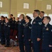 A group of United States Air Force personnel in dress blue uniforms are standing at attention in formation inside what appears to be a ceremonial hall or event venue. The service members are arranged in rows and appear to be participating in a formal military ceremony, possibly reciting an oath or singing. Both male and female airmen of various ranks are visible, identifiable by chevron insignia and service ribbons on their uniforms.