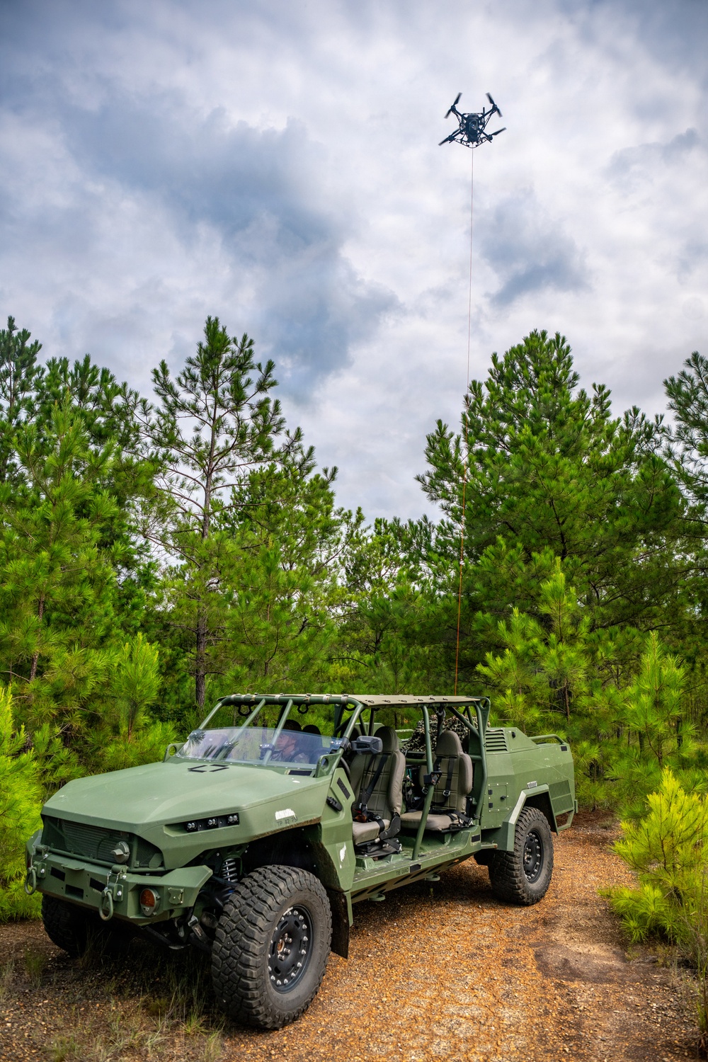 A military light tactical vehicle painted in olive drab green is parked on a dirt trail surrounded by pine trees. A multi-rotor drone (quadcopter or hexacopter) is tethered to the vehicle via a visible cable or tether line, hovering at significant altitude above. This appears to be a demonstration or operational test of a tethered drone system integrated with a ground vehicle platform.