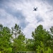 A military light tactical vehicle painted in olive drab green is parked on a dirt trail surrounded by pine trees. A multi-rotor drone (quadcopter or hexacopter) is tethered to the vehicle via a visible cable or tether line, hovering at significant altitude above. This appears to be a demonstration or operational test of a tethered drone system integrated with a ground vehicle platform.