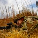 A soldier in full camouflage uniform is lying prone in a field of dry golden grass, aiming a rifle equipped with an optical scope. The soldier appears to be in a training or tactical exercise environment, with a target marker structure visible in the background. The image is taken from a low ground-level perspective, emphasizing the soldier's concealed firing position.