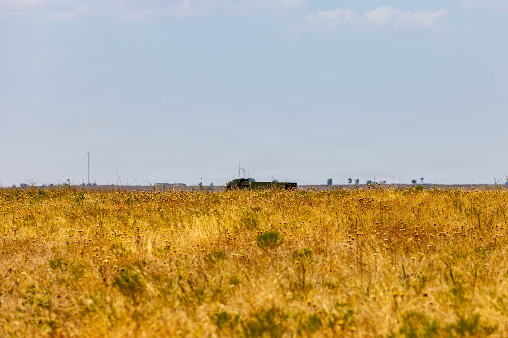 A wide open field of dry, golden-yellow grass or grain stretches across the foreground under a pale blue sky. In the far distance, a dark-colored military-style vehicle or heavy truck is partially visible above the vegetation line, along with what appear to be distant trees, structures, and a communications tower on the horizon. The scene suggests a flat, arid or semi-arid landscape, possibly a steppe or agricultural plain.