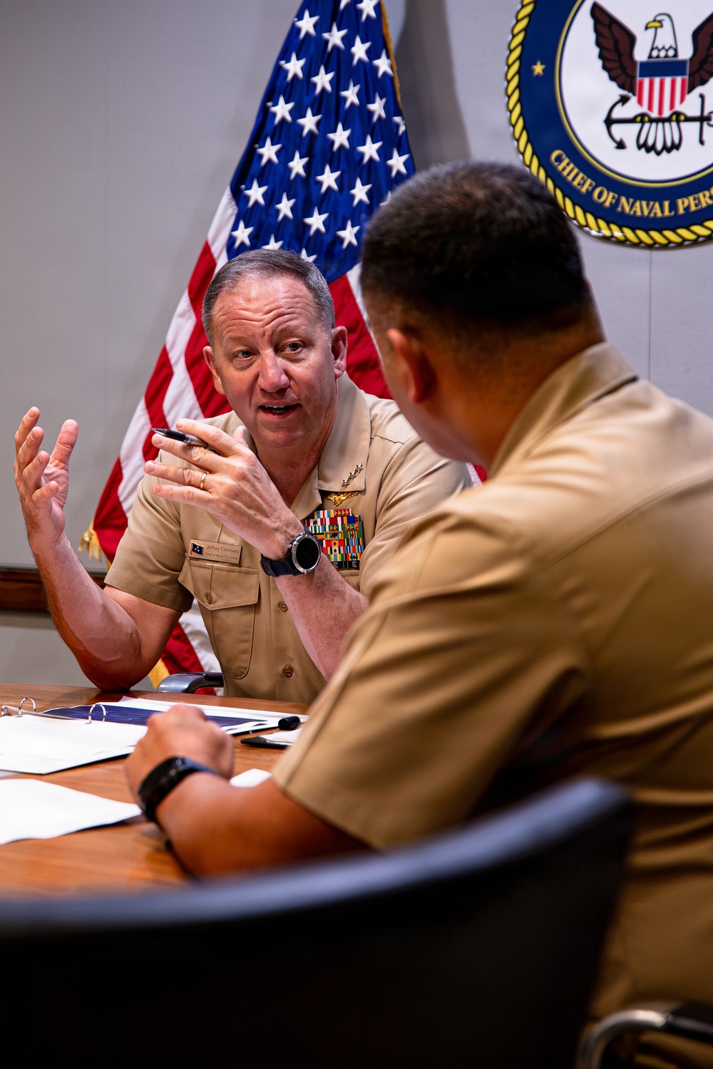 Two U.S. Navy officers in tan service uniforms are seated at a conference table engaged in discussion. The officer facing the camera is gesturing with both hands while speaking, with multiple service ribbons and a warfare device visible on his uniform. The setting is an official Navy office, indicated by the U.S. flag and a 'Chief of Naval Personnel' seal on the wall behind them.