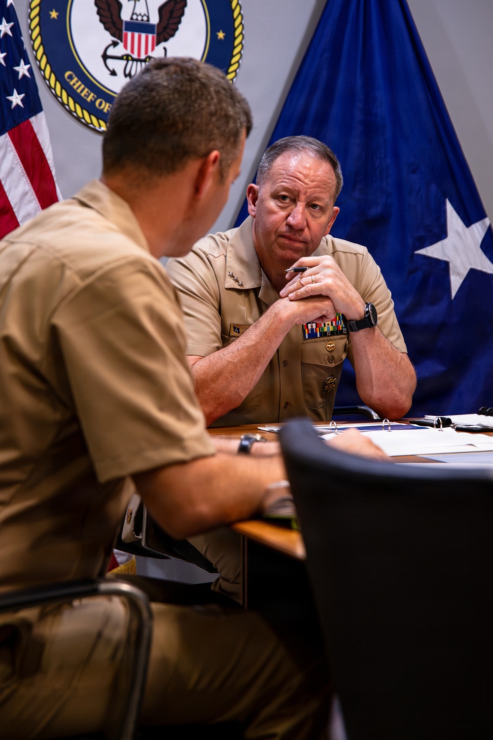Two U.S. Navy officers in tan service uniforms are engaged in a meeting at a conference table. The officer facing the camera appears to be a senior flag officer, visible by collar insignia and a blue flag with stars in the background. The setting is an official Navy office, indicated by the 'Chief of Naval Operations' seal on the wall.