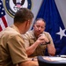 Two U.S. Navy officers in tan service uniforms are engaged in a meeting at a conference table. The officer facing the camera appears to be a senior flag officer, visible by collar insignia and a blue flag with stars in the background. The setting is an official Navy office, indicated by the 'Chief of Naval Operations' seal on the wall.