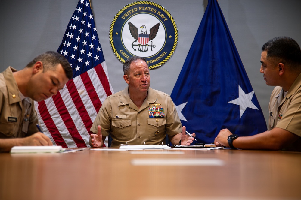 Three United States Navy personnel are seated at a conference table in what appears to be an official meeting setting. The central figure, a senior officer with multiple service ribbons and insignia, appears to be speaking or briefing the others. The setting is formal, backed by the U.S. flag, a Navy flag with a star, and the official seal of the United States Navy Chief of Naval Personnel.
