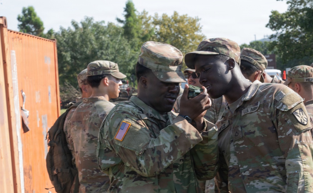 A group of U.S. Army soldiers in MultiCam/OCP camouflage uniforms are gathered outdoors near a shipping container. Two soldiers in the foreground appear to be examining a small object, possibly a compass or piece of equipment. The setting appears to be a training or staging area with trees and vehicles visible in the background.