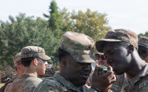 A group of U.S. Army soldiers in MultiCam/OCP camouflage uniforms are gathered outdoors near a shipping container. Two soldiers in the foreground appear to be examining a small object, possibly a compass or piece of equipment. The setting appears to be a training or staging area with trees and vehicles visible in the background.