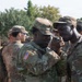 A group of U.S. Army soldiers in MultiCam/OCP camouflage uniforms are gathered outdoors near a shipping container. Two soldiers in the foreground appear to be examining a small object, possibly a compass or piece of equipment. The setting appears to be a training or staging area with trees and vehicles visible in the background.