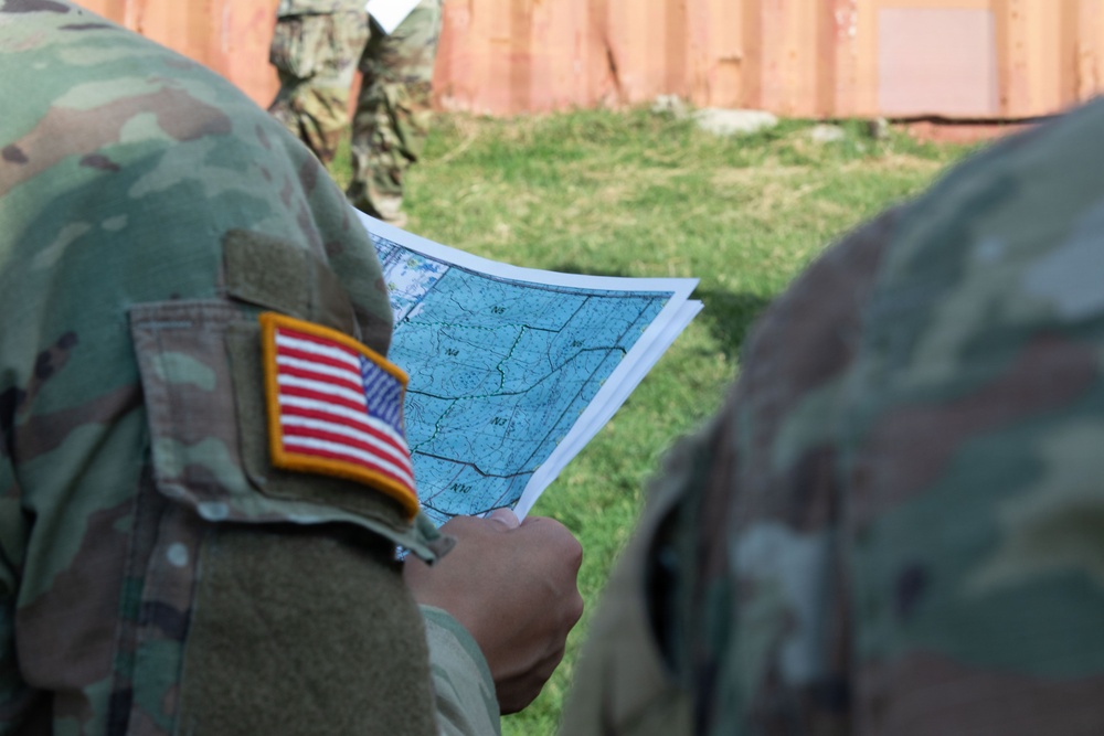 A U.S. Army soldier wearing Operational Camouflage Pattern (OCP) uniform with an American flag patch on the left shoulder is reviewing a topographic or tactical map. At least two soldiers are visible in the foreground, with a third soldier partially visible in the background near what appears to be a shipping container or metal wall. The setting appears to be an outdoor training or operational environment.