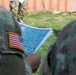A U.S. Army soldier wearing Operational Camouflage Pattern (OCP) uniform with an American flag patch on the left shoulder is reviewing a topographic or tactical map. At least two soldiers are visible in the foreground, with a third soldier partially visible in the background near what appears to be a shipping container or metal wall. The setting appears to be an outdoor training or operational environment.