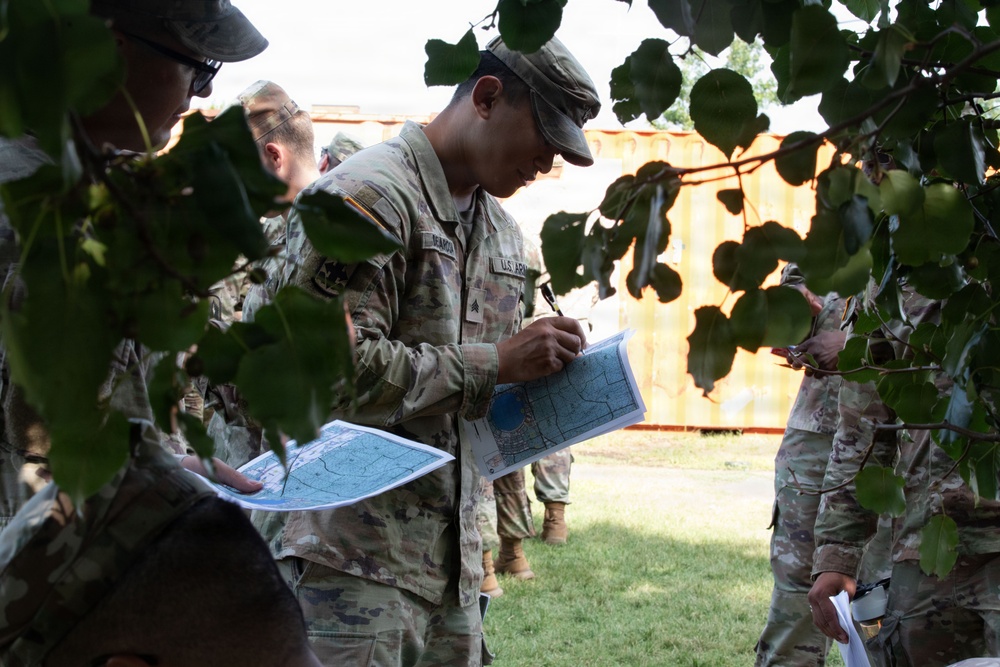 A group of U.S. Army soldiers in OCP (Operational Camouflage Pattern) uniforms are gathered outdoors, studying and annotating maps. The central figure, identifiable as a Sergeant based on visible rank insignia, is writing on a tactical map while others around him review similar documents. The setting appears to be a training or operational briefing area near a storage container.