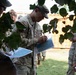 A group of U.S. Army soldiers in OCP (Operational Camouflage Pattern) uniforms are gathered outdoors, studying and annotating maps. The central figure, identifiable as a Sergeant based on visible rank insignia, is writing on a tactical map while others around him review similar documents. The setting appears to be a training or operational briefing area near a storage container.