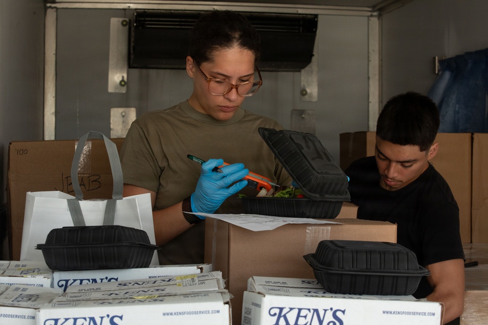 Two individuals are working inside what appears to be a refrigerated truck or storage trailer, handling food containers and boxes. The person in the foreground, wearing glasses, a olive/tan t-shirt, and blue latex gloves, is inspecting or labeling a black clamshell food container containing what appears to be a salad. Several branded 'Ken's Food Service' boxes are visible in the foreground along with multiple black takeout containers.