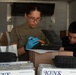 Two individuals are working inside what appears to be a refrigerated truck or storage trailer, handling food containers and boxes. The person in the foreground, wearing glasses, a olive/tan t-shirt, and blue latex gloves, is inspecting or labeling a black clamshell food container containing what appears to be a salad. Several branded 'Ken's Food Service' boxes are visible in the foreground along with multiple black takeout containers.