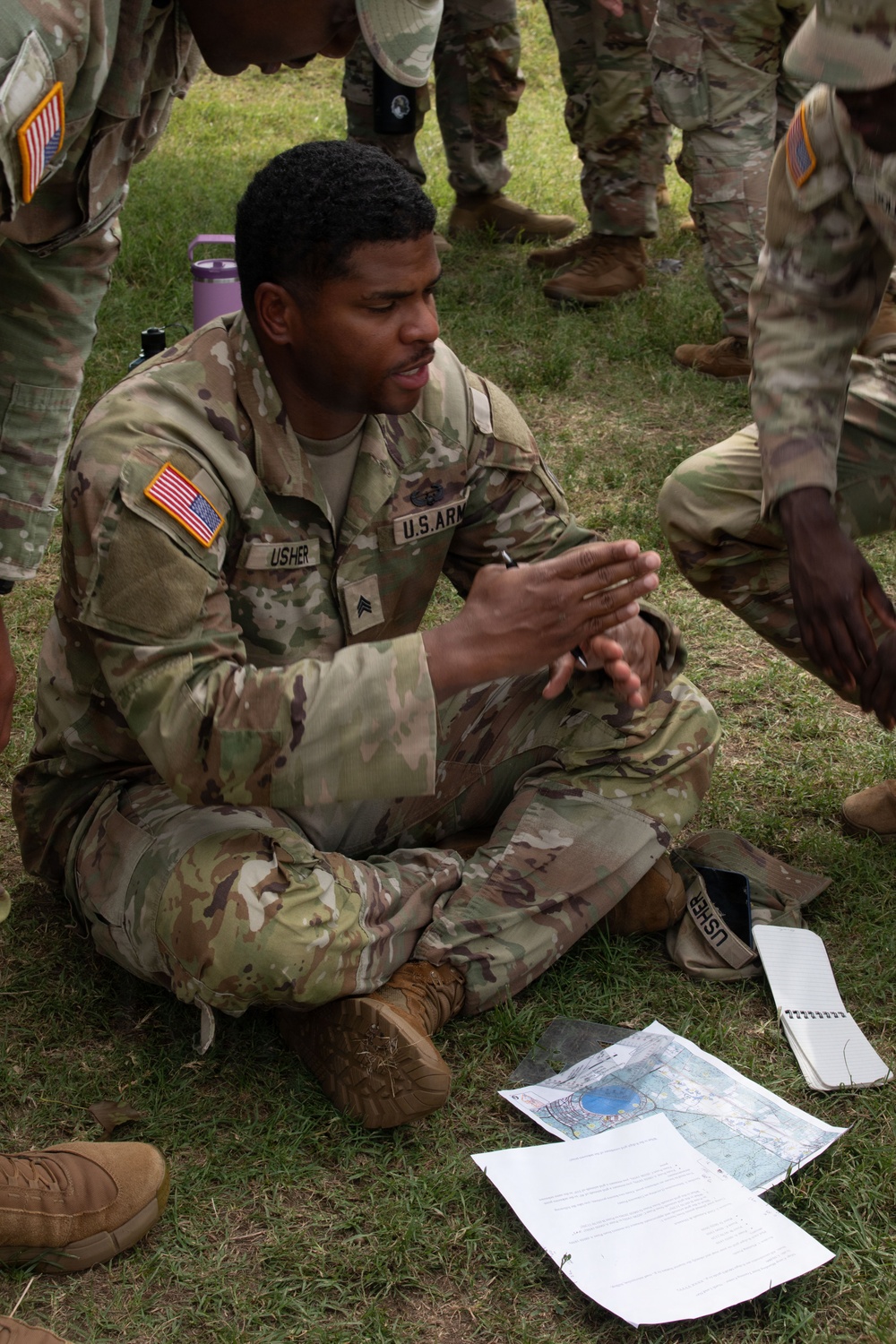 A U.S. Army Sergeant named Usher is seated cross-legged on grass, appearing to brief or instruct a group of fellow soldiers. He is gesturing with his hands while referencing documents including a topographic map and printed papers laid on the ground in front of him. The setting appears to be an outdoor field training exercise.