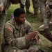 A U.S. Army Sergeant named Usher is seated cross-legged on grass, appearing to brief or instruct a group of fellow soldiers. He is gesturing with his hands while referencing documents including a topographic map and printed papers laid on the ground in front of him. The setting appears to be an outdoor field training exercise.