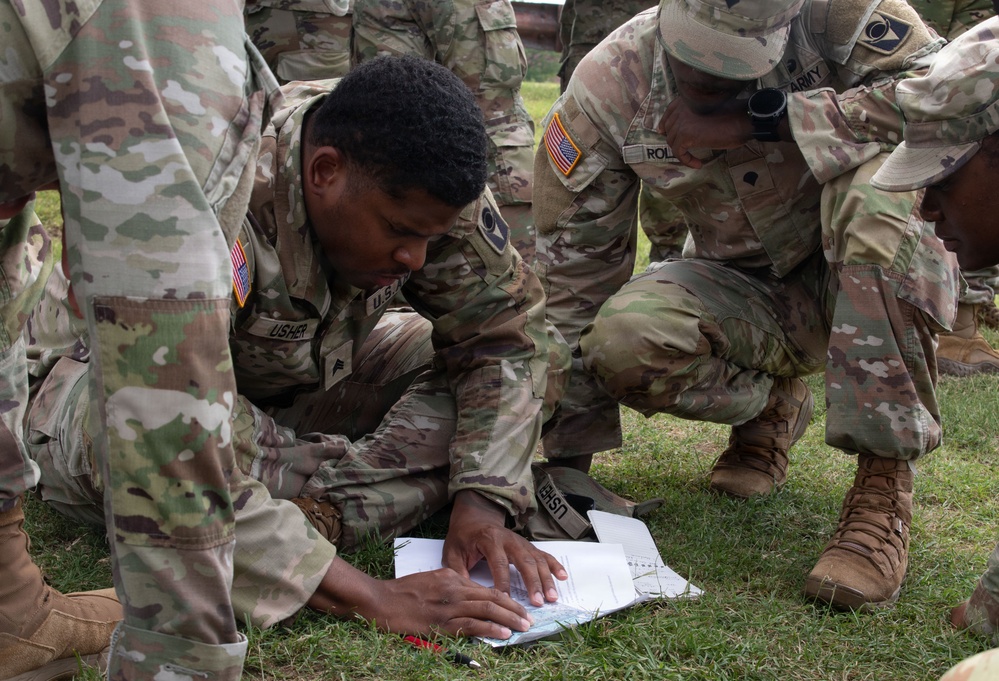 U.S. Army soldiers in Operational Camouflage Pattern (OCP) uniforms are gathered on a grassy field, studying documents or maps laid on the ground. The central soldier, identified by the name tape 'USHER,' is closely examining printed materials while fellow soldiers crouch around him. This appears to be a training exercise involving land navigation or tactical planning.