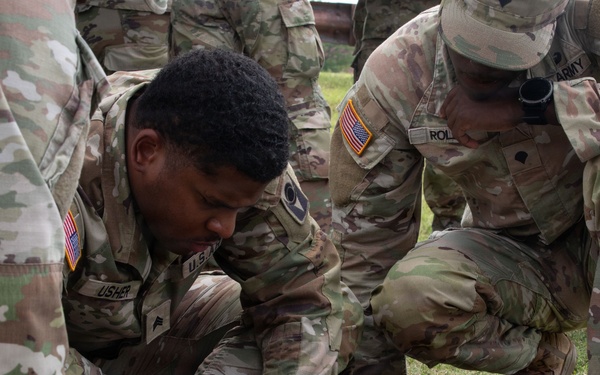 U.S. Army soldiers in Operational Camouflage Pattern (OCP) uniforms are gathered on a grassy field, studying documents or maps laid on the ground. The central soldier, identified by the name tape 'USHER,' is closely examining printed materials while fellow soldiers crouch around him. This appears to be a training exercise involving land navigation or tactical planning.