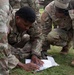 U.S. Army soldiers in Operational Camouflage Pattern (OCP) uniforms are gathered on a grassy field, studying documents or maps laid on the ground. The central soldier, identified by the name tape 'USHER,' is closely examining printed materials while fellow soldiers crouch around him. This appears to be a training exercise involving land navigation or tactical planning.