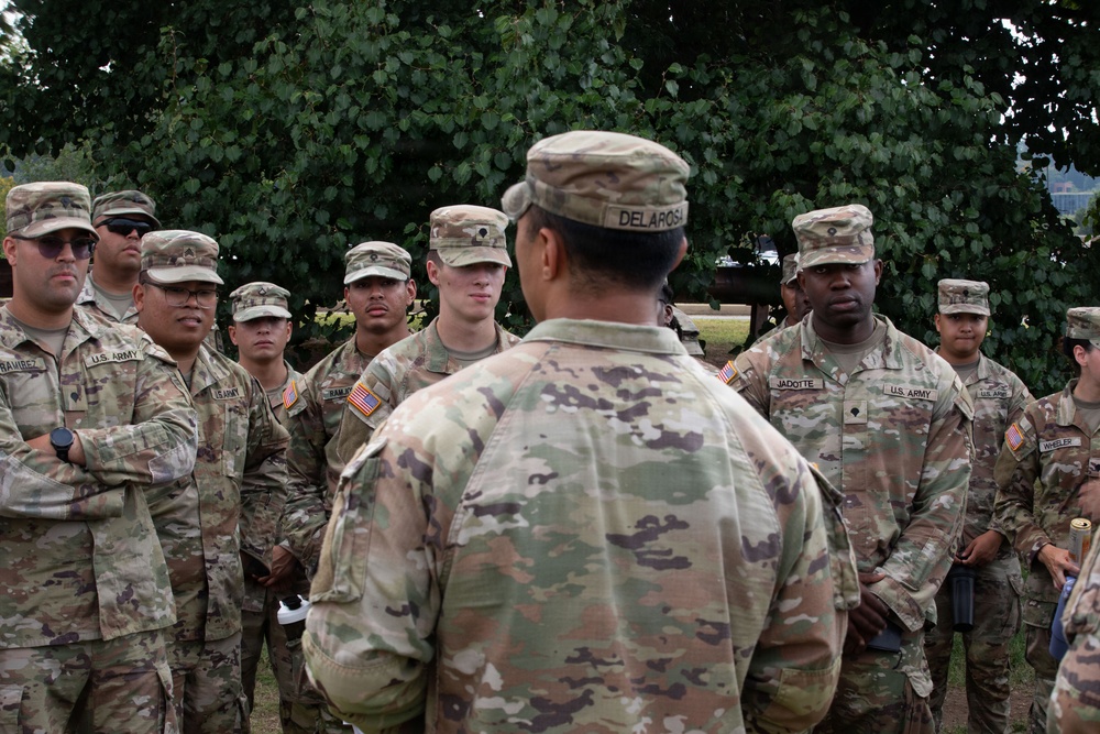 A U.S. Army soldier, identified by name tape as 'DELAROSA,' addresses a group of fellow soldiers in an outdoor setting with trees in the background. The soldiers are wearing Army Combat Uniforms (ACU) in OCP (Operational Camouflage Pattern) and patrol caps. This appears to be a briefing or formation gathering, with visible name tapes including JADOTTE, WHEELER, RAMIREZ, and RAMIREZ among others.