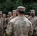 A U.S. Army soldier, identified by name tape as 'DELAROSA,' addresses a group of fellow soldiers in an outdoor setting with trees in the background. The soldiers are wearing Army Combat Uniforms (ACU) in OCP (Operational Camouflage Pattern) and patrol caps. This appears to be a briefing or formation gathering, with visible name tapes including JADOTTE, WHEELER, RAMIREZ, and RAMIREZ among others.