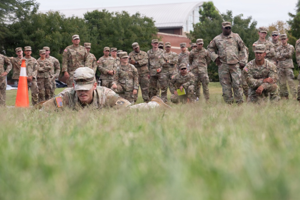 A group of U.S. Army soldiers in MultiCam/OCP camouflage uniforms are gathered on a grassy field, observing a fellow soldier performing a low crawl exercise. The soldier in the foreground is prone on the grass, executing what appears to be a field movement technique demonstration or evaluation. A large group of soldiers stands watching in the background, suggesting a training or assessment event.