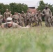 A group of U.S. Army soldiers in MultiCam/OCP camouflage uniforms are gathered on a grassy field, observing a fellow soldier performing a low crawl exercise. The soldier in the foreground is prone on the grass, executing what appears to be a field movement technique demonstration or evaluation. A large group of soldiers stands watching in the background, suggesting a training or assessment event.