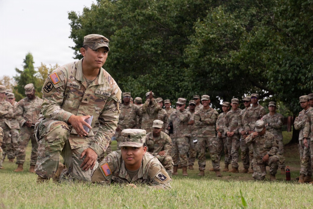 A U.S. Army training exercise is taking place outdoors on a grassy field. A soldier identified by nametape as 'DELAROSA' is kneeling and supervising another soldier who is in a prone position on the ground. A large group of uniformed soldiers observes in the background, suggesting this is a demonstration or evaluation event.
