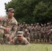 A U.S. Army training exercise is taking place outdoors on a grassy field. A soldier identified by nametape as 'DELAROSA' is kneeling and supervising another soldier who is in a prone position on the ground. A large group of uniformed soldiers observes in the background, suggesting this is a demonstration or evaluation event.
