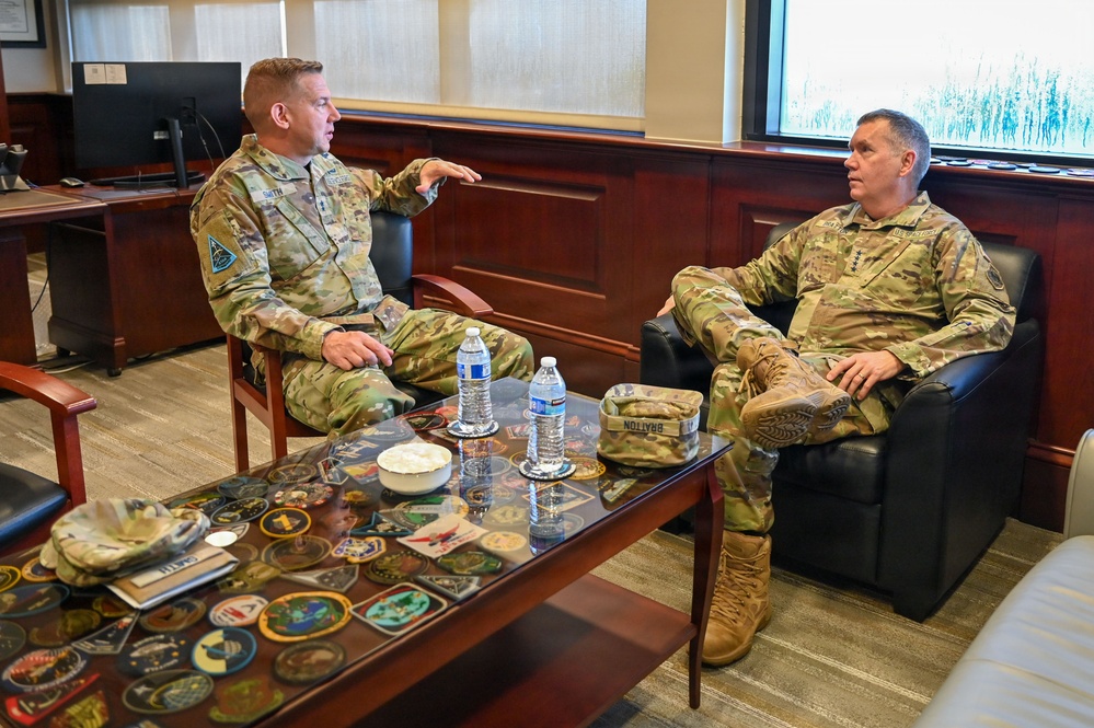 Two military personnel in U.S. Army OCP (Operational Camouflage Pattern) uniforms are seated in a formal office setting, engaged in conversation. The individual on the left appears to be speaking and gesturing, while the one on the right listens attentively. A glass-topped coffee table displaying military unit patches and challenge coins is visible between them.