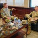 Two military personnel in U.S. Army OCP (Operational Camouflage Pattern) uniforms are seated in a formal office setting, engaged in conversation. The individual on the left appears to be speaking and gesturing, while the one on the right listens attentively. A glass-topped coffee table displaying military unit patches and challenge coins is visible between them.