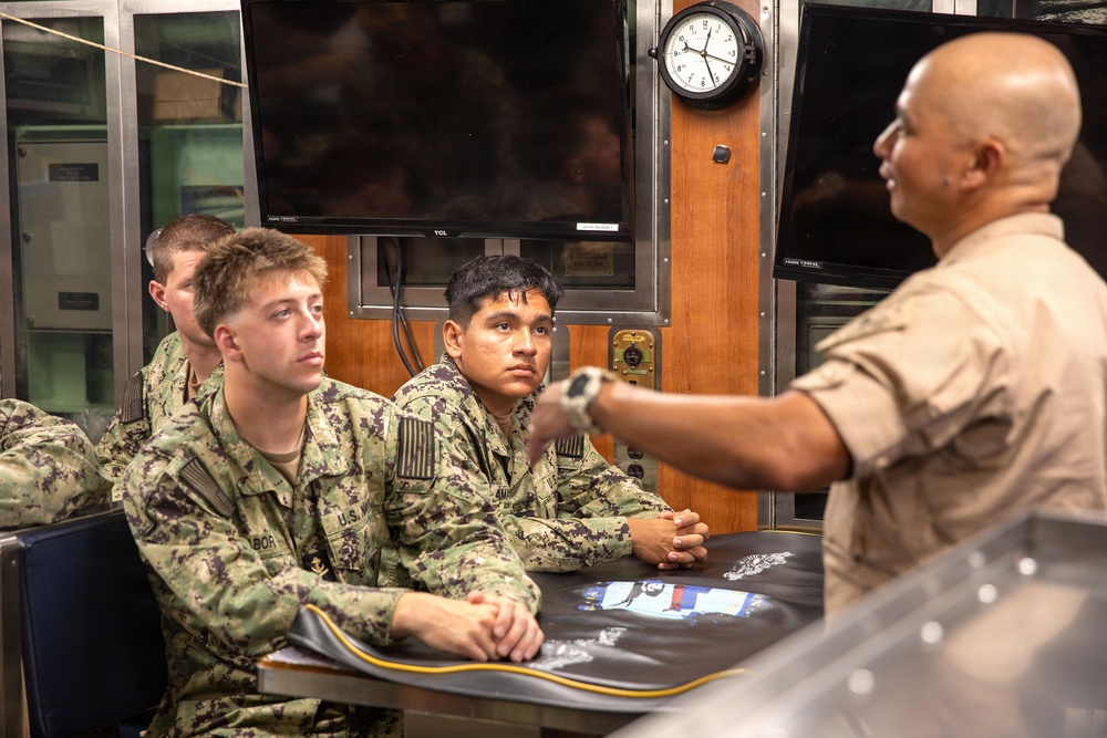 A military briefing or instruction session is taking place in what appears to be a naval operations room or ship interior. Several personnel in U.S. Navy digital camouflage uniforms are seated at a table listening attentively to an instructor or senior personnel wearing a tan/khaki uniform. The setting includes multiple monitors, a wall clock, and wooden panel fixtures consistent with a naval vessel or facility.