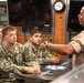 A military briefing or instruction session is taking place in what appears to be a naval operations room or ship interior. Several personnel in U.S. Navy digital camouflage uniforms are seated at a table listening attentively to an instructor or senior personnel wearing a tan/khaki uniform. The setting includes multiple monitors, a wall clock, and wooden panel fixtures consistent with a naval vessel or facility.