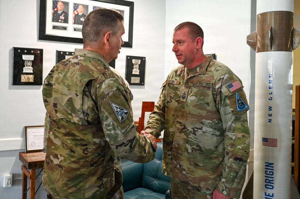 Two U.S. Space Force personnel in OCP (Operational Camouflage Pattern) uniforms are shaking hands in what appears to be an office or reception area. A scale model of Blue Origin's New Glenn rocket is prominently displayed to the right. The setting suggests an official military visit or recognition ceremony.