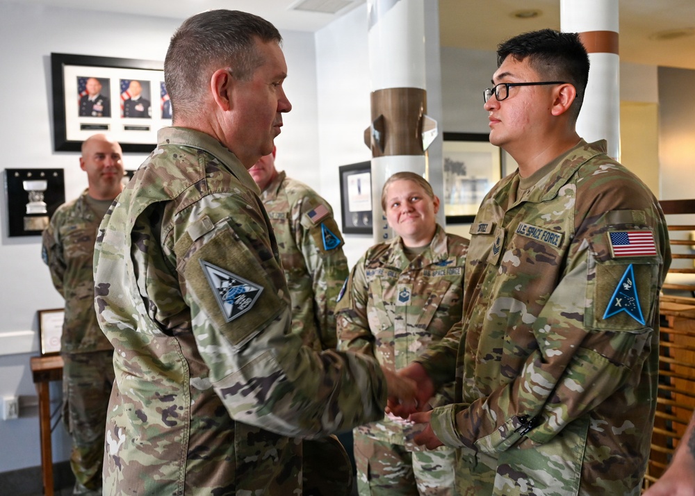 A military ceremony or recognition event is taking place indoors, where a senior officer is shaking hands with a U.S. Space Force member, likely presenting a coin or award. Several other uniformed personnel are present in the background, observing the exchange. The setting appears to be an official military building with framed portraits and display cases on the walls.