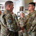 A military ceremony or recognition event is taking place indoors, where a senior officer is shaking hands with a U.S. Space Force member, likely presenting a coin or award. Several other uniformed personnel are present in the background, observing the exchange. The setting appears to be an official military building with framed portraits and display cases on the walls.