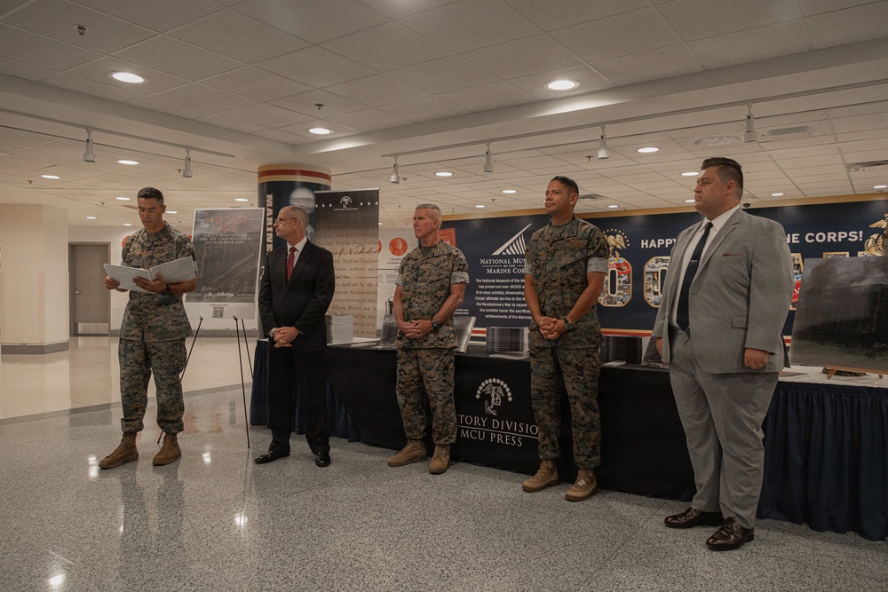 A formal military ceremony or presentation is taking place in what appears to be a government or military building. Five individuals are standing in a line — three in U.S. Marine Corps combat utility uniforms and two in civilian business attire. The backdrop includes displays referencing the National Museum of the Marine Corps, MCCU Press History Division, and a banner reading 'Happy [Birthday] Marine Corps.'