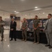 A formal military ceremony or presentation is taking place in what appears to be a government or military building. Five individuals are standing in a line — three in U.S. Marine Corps combat utility uniforms and two in civilian business attire. The backdrop includes displays referencing the National Museum of the Marine Corps, MCCU Press History Division, and a banner reading 'Happy [Birthday] Marine Corps.'